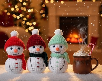 May include: Three crocheted snowmen wearing red, burgundy, and mint green hats and scarves. Each snowman has a carrot nose and a smiling face. A mug of hot chocolate with whipped cream and a candy cane sits to the right.