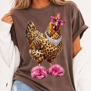 Pode incluir: Uma t-shirt castanha de manga curta com o gráfico de uma galinha com penas de padrão leopardo, usando óculos de sol cat-eye cor-de-rosa brilhante, um colar de strass prateado e chinelos cor-de-rosa felpudos nos pés.
