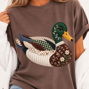 Könnte beinhalten: Ein braunes T-Shirt mit kurzen Ärmeln und einer großen Entengrafik, die aus verschiedenen Stoffmustern zusammengesetzt ist. Das Entendesign umfasst grüne, braune und cremefarbene Bereiche mit Blumen-, Vichy- und Streifenmustern sowie weißen Gänseblümchen.