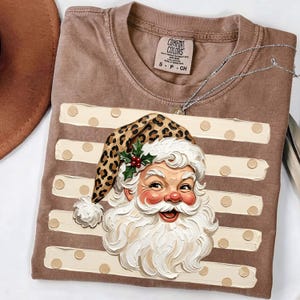 May include: A brown t-shirt featuring a graphic of Santa Claus wearing a leopard print hat with a holly sprig. The background of the graphic consists of horizontal cream-coloured stripes with polka dots. The shirt has a Comfort Colors brand label.