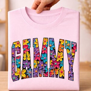 May include: A light pink t-shirt featuring the word GAMMY in large, block-style letters. The interior of each letter is filled with a vibrant, multi-colored pattern of small, textured flowers in shades of yellow, pink, orange, blue, and purple.