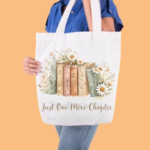 Puede incluir: Bolso tote blanco con un diseño de acuarela de libros y margaritas. Los libros son en tonos verdes, beige y marrón. El texto "Just One More Chapter" está impreso en una fuente cursiva en la parte inferior de la bolsa.