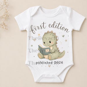 May include: A white short-sleeved baby bodysuit featuring a graphic of a light green dinosaur reading a book. The text reads first edition above the dinosaur and published 2026 below it, surrounded by small stars and hearts in soft neutral tones.