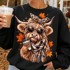 May include: A black long-sleeve sweatshirt featuring a graphic of a fluffy Highland cow wearing leopard print sunglasses and a matching bow. The cow has a plaid scarf, holds a coffee cup, and is surrounded by orange autumn maple leaves.