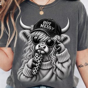May include: A charcoal gray t-shirt featuring a graphic of a shaggy highland cow wearing a black baseball cap with the text HOT MESS and a heart symbol. The cow wears leopard print sunglasses and holds a matching leopard print tumbler cup.