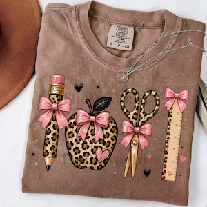 May include: A brown t-shirt with a Comfort Colors label featuring a graphic print of a pencil, an apple, scissors, and a ruler, all decorated with a leopard print pattern and light pink bows, accented by small black hearts and gold stars.