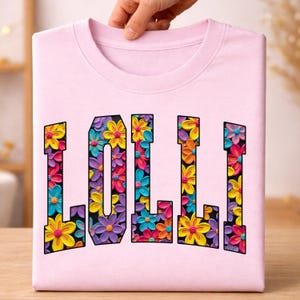 Peut inclure: Un t-shirt rose clair avec le mot LOLLI écrit en grandes lettres capitales. L'intérieur de chaque lettre est rempli d'un motif texturé et vibrant de fleurs colorées dans des tons jaune, rose, bleu et orange sur un fond noir.