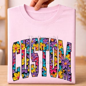May include: A light pink t-shirt featuring the word CUSTOM in large block letters. The interior of each letter is filled with a dense, textured pattern of small, colorful flowers in shades of yellow, purple, orange, and blue on a black background.