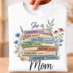 Könnte beinhalten: Weißes Sweatshirt mit einem Design aus gestapelten Büchern und Blumen. Jedes Buch zeigt ein positives Wort und einen Bibelverweis. Die Worte "She is Mom" sind in einer Schriftart geschrieben.