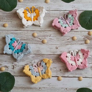 Könnte beinhalten: Eine Sammlung von fünf handgefertigten Häkel-Schmetterlingsapplikationen in verschiedenen Farben, darunter Weiß, Gelb, Rosa, Blau und Grau. Die Schmetterlinge sind auf einer hellen Holzoberfläche angeordnet, mit kleinen Holzperlen und grünen Blättern.