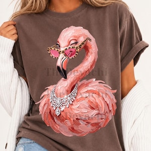 Puede incluir: Una camiseta marrón de manga corta con el gráfico de un flamenco rosa que lleva gafas con estampado de leopardo y un collar de pedrería plateado. El flamenco guiña un ojo y está representado con un estilo pictórico texturizado y pinceladas visibles.