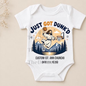 Op de afbeelding: Een witte babybody met korte mouwen en een afbeelding van een figuur die een basketbal dunt, met de tekst JUST GOT DUNK'D, CUSTOM (ST. ANN CHURCH) en DATE (11.10.26). Het ontwerp bevat bergen, dennenbomen en sterren.
