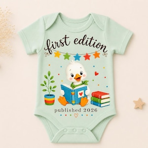 May include: A light green short-sleeve baby bodysuit featuring a graphic of a white duckling reading a blue book. The text reads first edition and published 2026. The design includes a colorful bunting, a potted plant, and a stack of three books.
