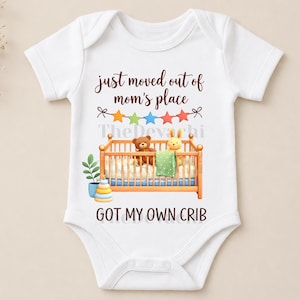 May include: White baby onesie with the text "just moved out of mom's place" and "got my own crib". The design includes a crib with a teddy bear and a bunny. A colorful star banner is above the crib.