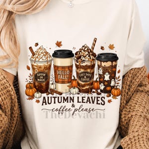 Könnte beinhalten: Ein cremefarbenes T-Shirt mit einer Grafik von vier Kaffeetassen, verziert mit herbstlichen Motiven wie Kürbissen, Blättern und Geistern. Der Text lautet Sippin' on Magic, Hello Fall, Cozy Days & Coffee und Autumn Leaves & coffee please.