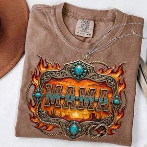 May include: A brown short-sleeve t-shirt featuring a graphic print of a decorative metal frame with turquoise accents, flames, and a desert landscape. The text M-A-M-A is centred within the frame. A silver chain necklace rests on the shirt.