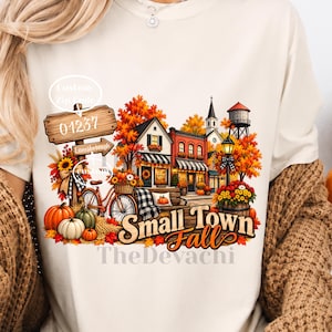 May include: A cream-colored t-shirt featuring a graphic of a small town in autumn with orange trees, pumpkins, a bicycle, and a wooden signpost labeled 01237 and Lanesborough. The text Small Town Fall is printed in large, stylized orange and brown letters.