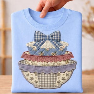 Könnte beinhalten: Hellblaues Sweatshirt mit einem Design aus gestapelten dekorativen Schalen und einer Schleife. Die Schalen haben Blumen- und Vichy-Muster in Blau, Rosa und Lila. Das Design wird von einer Hand gehalten.