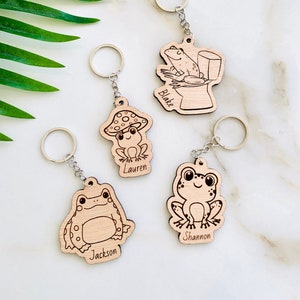 Peut inclure: Quatre porte-clés en bois avec des motifs de grenouilles. Chaque porte-clés a un motif de grenouille différent et un nom gravé en bas. Les noms sont Blake, Lauren, Jackson et Shannon.