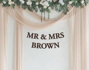 May include: A wedding backdrop featuring a white wall, sheer beige curtains, and a greenery garland with white flowers. The text "MR & MRS BROWN" is displayed in brown letters on the wall.