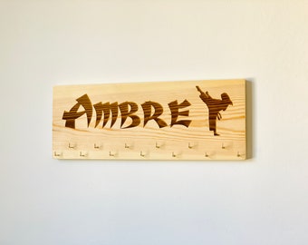 Könnte beinhalten: Ein Holzmedaillenhalter mit dem Namen "AMBREY" in einer stilisierten Schriftart graviert. Eine Silhouette einer Person in einer Karate-Pose ist ebenfalls auf dem Halter graviert. Es gibt 10 Haken an der Unterseite des Halters zum Aufhängen von Medaillen.