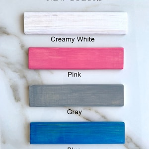 May include: Four wooden samples painted in different colors: creamy white, pink, gray, and blue. The text "NEW COLORS" is at the top of the image.