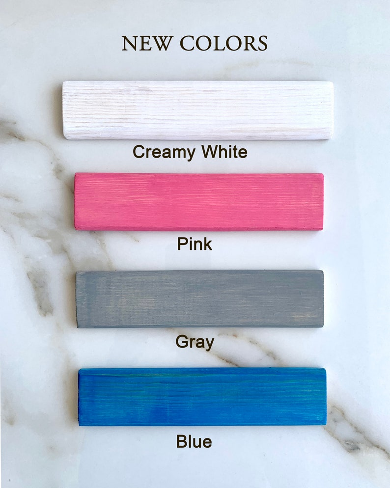 May include: Four wooden samples painted in different colors: creamy white, pink, gray, and blue. The text "NEW COLORS" is at the top of the image.
