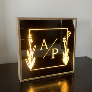 May include: A wooden framed light-up sign with a black background and a gold square frame. The initials "A/P" are in the center of the square with a floral design on each side.