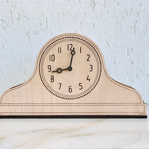 May include: A wooden clock with a light brown finish and black numbers. The clock has a simple design with a round face and a rectangular base. The clock is set to 12:00.
