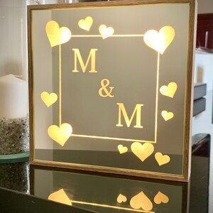 May include: A wooden framed mirror with a gold-colored inscription that reads "M & M" surrounded by small hearts. The mirror is illuminated from behind, creating a warm glow.