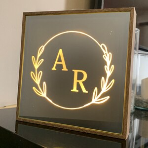 May include: A wooden framed mirror with a gold-colored background and a lit-up circle with the letters "A" and "R" inside. The circle is surrounded by a wreath of leaves.