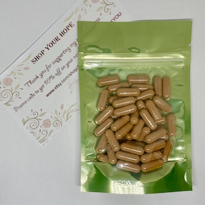 May include: A green resealable pouch filled with approximately 30 brown capsules. The pouch has a zipper closure and is made of a shiny, metallic material. A small card with the text "SHOP YOUR HOPE" is visible in the background.