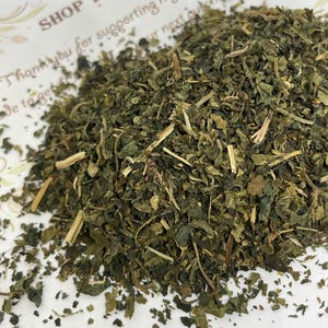 May include: A close-up of a pile of dried green nettle leaves. The leaves are finely chopped and have a dark green color.