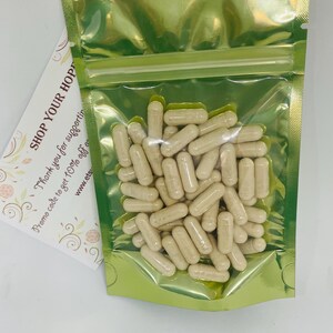 May include: A clear plastic bag filled with beige capsules. The bag is sealed with a gold-colored zipper and is set against a white background. A card with the text "SHOP YOUR HOP" is visible.