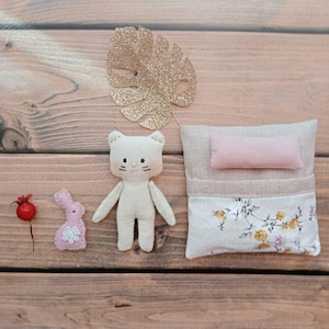 Könnte beinhalten: Eine Sammlung handgefertigter Stoffspielzeuge und Dekorationen auf einer Holzoberfläche. Enthält eine cremefarbene Katzendoll, ein rosa Häschen mit floralem Design, ein goldglitzerndes Blatt, ein kleines Kissen und ein Bett mit floralen Akzenten.