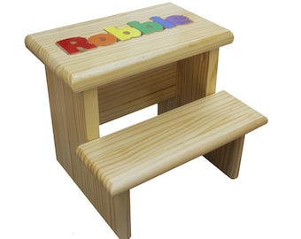 Puede incluir: Un taburete de madera clara con dos escalones. El nombre "Robbie" está escrito con letras coloridas en el escalón superior. El taburete es de madera natural y tiene un diseño simple y resistente, ideal para alcanzar lugares más altos.