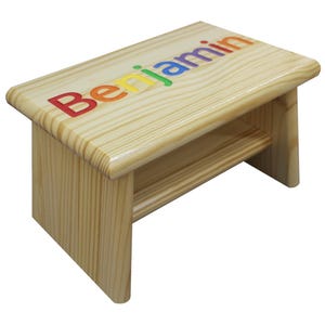 May include: A small, wooden step stool with the name "Benjamin" printed across the top in rainbow colors. The stool is made of light-colored wood and has a simple, rectangular design with rounded edges.