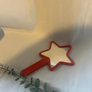 Peut inclure: Un miroir de poche rouge en forme d'étoile avec un manche. Le miroir est sur une surface blanche avec une tige verte feuillue.