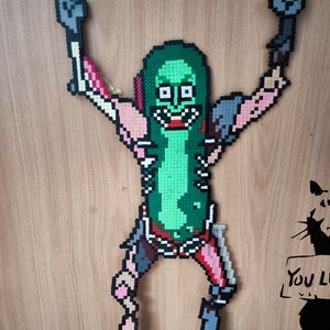 Op de afbeelding: Een groen augurk personage met een skeletlichaam en armen omhoog in de lucht. Het personage is gemaakt van kleurrijke plastic kralen.