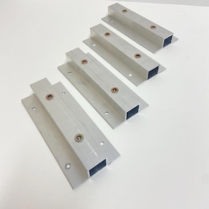 May include: Four silver-colored metal brackets, each featuring a rectangular hollow profile attached to a flat base plate. Each bracket includes two circular openings with metallic hardware inserts, designed for mounting purposes.