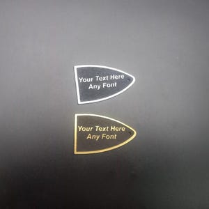 Peut inclure: Deux plaques noires en forme de médiator de guitare avec une bordure dorée. La plaque du haut a du texte blanc qui dit "Your Text Here Any Font". La plaque du bas a du texte doré qui dit "Your Text Here Any Font".