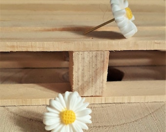 May include: Two white daisy earrings with yellow centers. The earrings are on a wooden surface.