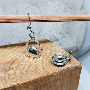 Può includere: Un paio di orecchini d'argento, uno con una teiera in miniatura e l'altro con una tazzina da tè in miniatura e un piattino. Il piattino ha incisa la scritta "Let's Get Saucy".