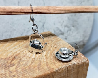 May include: A pair of silver earrings, one featuring a miniature teapot and the other a miniature teacup and saucer. The saucer has the text "Let's Get Saucy" engraved on it.