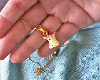 May include: A gold-colored necklace featuring a pendant shaped like an apple core. The pendant is white with red accents and a small black detail. The necklace is displayed against a light blue background.