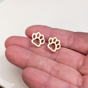 Puede incluir: Un par de pendientes de botón de color dorado con forma de huella de pata. Los pendientes son pequeños y delicados, y serían un gran regalo para cualquier amante de los perros.
