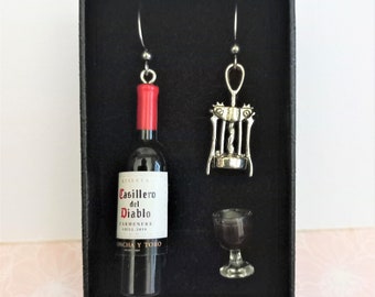 May include: A set of three silver wine-themed earrings in a black box. The earrings are a wine bottle, a corkscrew, and a wine glass. The wine bottle is labeled "Casillero del Diablo Carmenere Chile 2019 Concha y Toro."