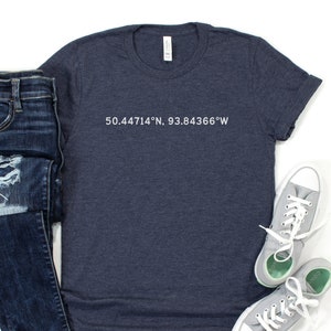 Peut inclure: Un t-shirt bleu marine avec un texte blanc qui dit "50.44714°N, 93.84366°W". Le t-shirt est posé à plat sur une surface blanche avec une paire de baskets grises et un jean bleu.
