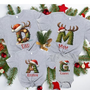 May include: A set of four grey t-shirts with Christmas-themed designs. The shirts feature the letters D, M, A, and G, each decorated with a Christmas tree, reindeer antlers, and a Santa hat. The shirts are personalised with the names "Dad", "Mom", "Addison", and "Gabriel".