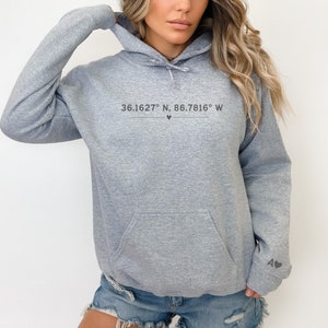 May include: A light gray hoodie with a front pocket and a hood. The text "36.1627° N, 86.7816° W" is printed on the chest. The left sleeve has the letters "A" and a heart symbol.